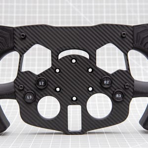 May include: A black racing wheel with a carbon fibre design. The wheel has buttons labelled L2, L3, R2, and R3. The grips have a textured pattern for enhanced grip.