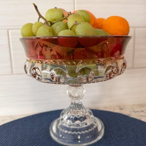 May include: A clear glass footed fruit bowl with a red rim and gold trim, filled with green grapes and oranges. The bowl has a decorative, patterned design and sits on a blue surface.