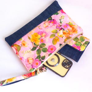 May include: A floral print clutch and matching cardholder with denim accents. The clutch features a pink background with yellow, pink, and orange flowers. A gold zipper and wrist strap complete the design. A yellow phone with a black case is inside the clutch.