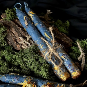 Puede incluir: Dos velas azules con flores secas y detalles dorados est&aacute;n atadas con cuerda y colocadas sobre un lecho de musgo verde.