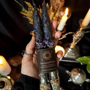 Könnte beinhalten: Zwei schwarze Ritualkerzen, verziert mit getrockneten blauen Blüten und goldenen Akzenten. Die Kerzen haben ein Etikett mit der Aufschrift "Ancestral Flame" mit Text über Schutz und Rituale. Die Kerzen sind handgefertigt.