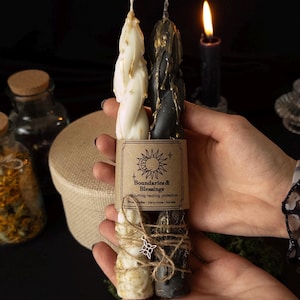 May include: Two twisted taper candles, one white and one black, both featuring gold wax drips. A brown paper label reads Boundaries & Blessings, cord cutting, healing, protection. The candles are tied with twine and a small silver metal charm.
