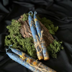 Puede incluir: Tres velas azules con detalles amarillos y dorados est&aacute;n atadas con cuerda. Las velas est&aacute;n sobre un lecho de musgo verde y un trozo de corteza de &aacute;rbol.