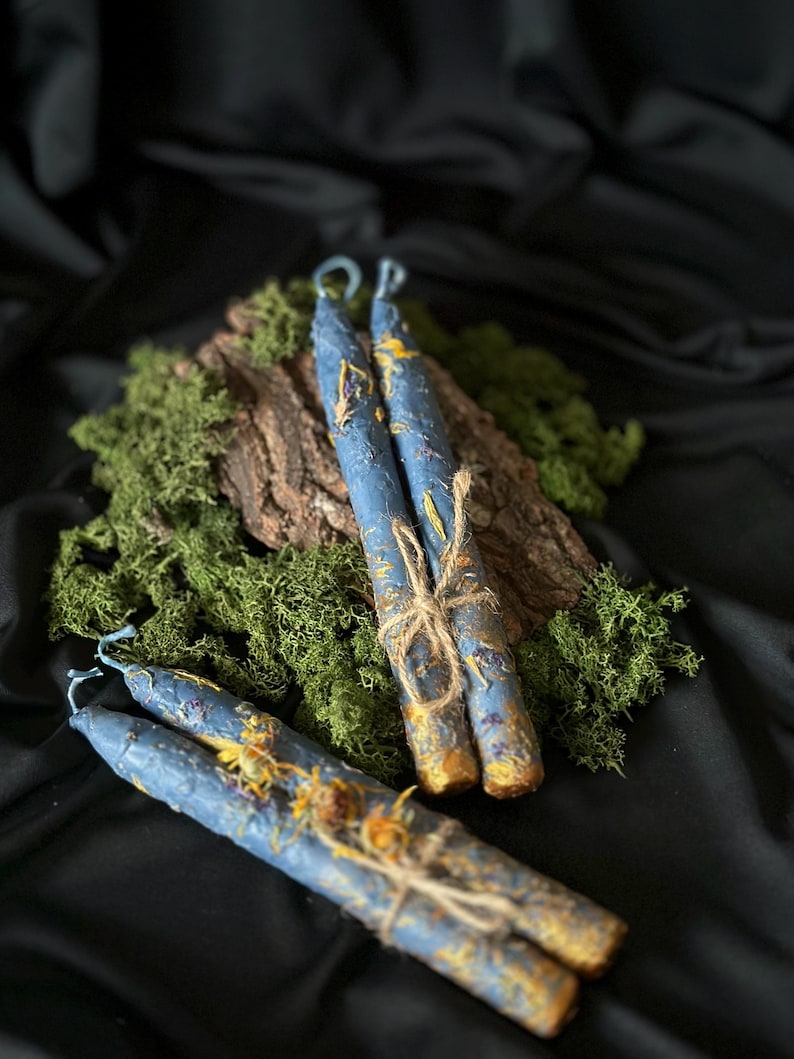 Puede incluir: Tres velas azules con detalles amarillos y dorados est&aacute;n atadas con cuerda. Las velas est&aacute;n sobre un lecho de musgo verde y un trozo de corteza de &aacute;rbol.