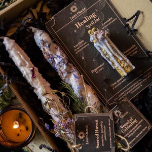 May include: A Light Oracle healing kit featuring candles, a spell jar, and a candle. The kit includes two white candles decorated with dried flowers, a glass jar with herbs, and a lit candle in a glass container. The kit is in a brown box with black paper.