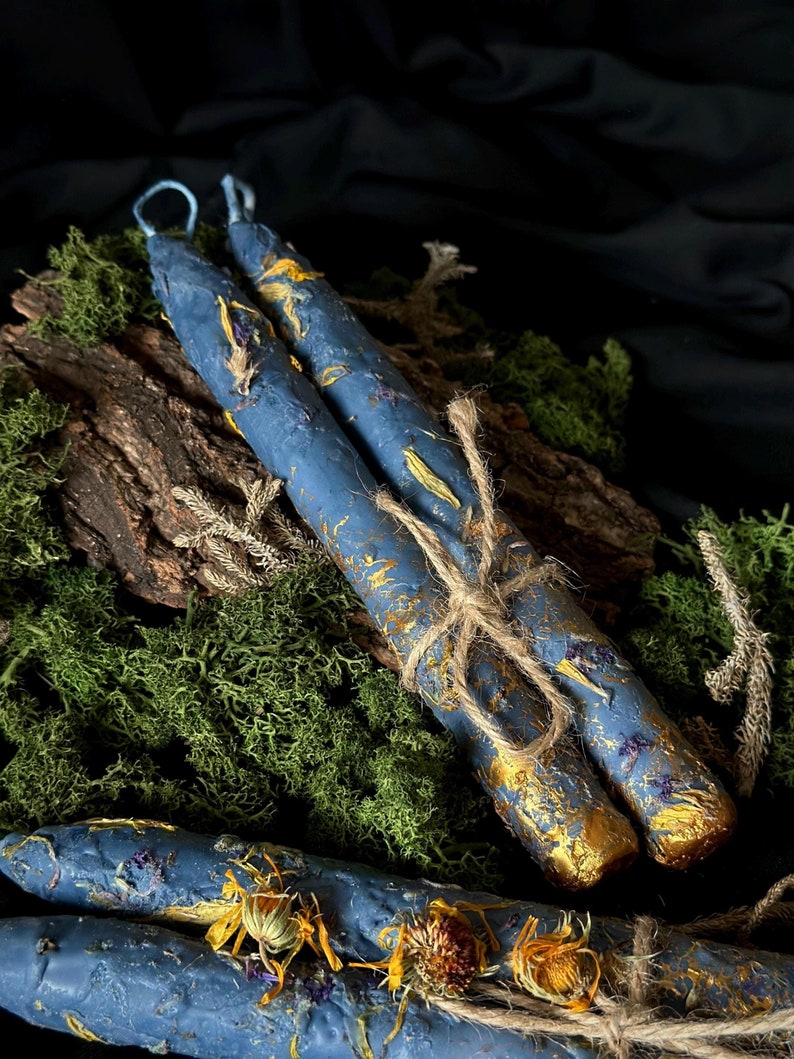 Puede incluir: Dos velas azules con flores secas y detalles dorados est&aacute;n atadas con cuerda y colocadas sobre un lecho de musgo verde.
