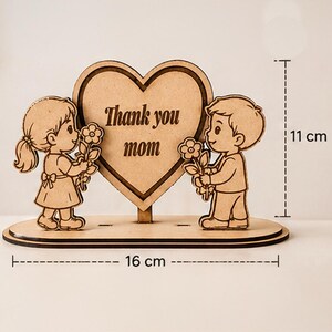 Op de afbeelding: Houten decoratiestuk met een hartvormig middelpunt met de tekst "Thank you mom". Twee cartoonfiguren, een meisje en een jongen, flankeren het hart, elk met een bloem. De basis is 16 cm lang en het stuk is 11 cm hoog.