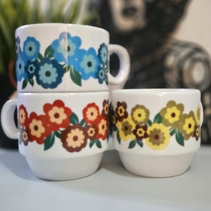 Könnte beinhalten: Drei weiße Keramikbecher, die übereinander gestapelt sind, jeder mit einem Blumendekor in Blau-, Rot- und Gelbtönen. Die Becher haben ein schlichtes Design mit Henkel und rundem Boden. Das Blumenmuster wiederholt sich am Becherkörper.