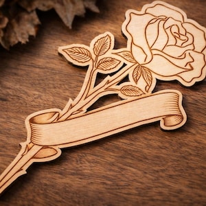 Puede incluir: Una rosa de madera con tallo, hojas y una cinta para texto. La rosa y las hojas están detalladas, y la madera tiene un tono marrón claro. La cinta está en blanco, lista para personalizar. Sobre una superficie de madera.