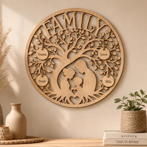 Puede incluir: Una pieza de arte mural de árbol genealógico de madera. La palabra "FAMILY" está recortada en la parte superior. El diseño del árbol incluye un corazón en la base y cuatro etiquetas con nombres: Emma, Noah, Luca y Mila. El arte es circular y de madera clara.