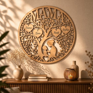 Op de afbeelding: Ronde houten wanddecoratie met het woord "MAMA" bovenaan uitgesneden. Een boomontwerp met hartvormige bladeren met de namen: Emma, Liam, Noah en Sophie. De boom bevat een moeder-kind silhouet. Gemaakt van lichtbruin hout.