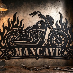 May include: Black metal motorcycle wall art with a skull on the engine, surrounded by flames. The word "MANCAVE" is displayed below the motorcycle. The art is mounted on a textured wall, with a vintage lamp above.