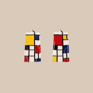 Può includere: Un paio di orecchini di perline con un design ispirato a Mondrian. Gli orecchini hanno una forma rettangolare con un motivo a griglia di perline rosse, gialle, blu e nere. Frange bianche pendono dal basso. Gli orecchini sono sospesi a ganci color argento.