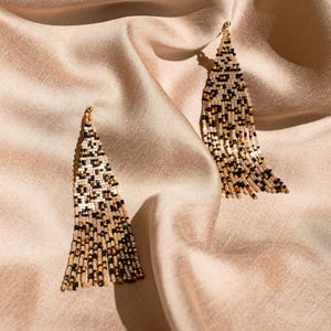May include: Two beaded earrings with a leopard print design. The earrings are made of small beads in black, tan, and gold colors. They hang from a gold-colored hook and are displayed on a beige satin fabric.