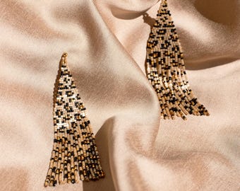 May include: Two beaded earrings with a leopard print design. The earrings are made of small beads in black, tan, and gold colors. They hang from a gold-colored hook and are displayed on a beige satin fabric.