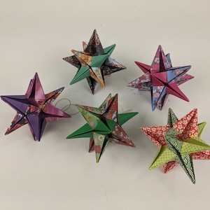 May include: Six origami stars in various colors and patterns. The stars are made of folded paper with floral and geometric designs. Each star has multiple points and a three-dimensional shape, suitable for holiday decor.