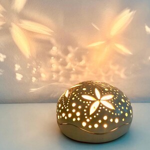 May include: A beige ceramic candle holder with a domed shape, featuring cut-out star and dot patterns. The candle holder casts star-shaped light patterns on the wall. The overall theme is home decor.