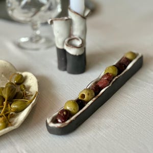 Puede incluir: Un plato para aceitunas de cerámica largo, estrecho y rectangular con exterior negro e interior blanco, lleno de una mezcla de aceitunas verdes y moradas oscuras. El plato presenta un borde ondulado y orgánico, hecho de arcilla oscura con esmalte blanco.