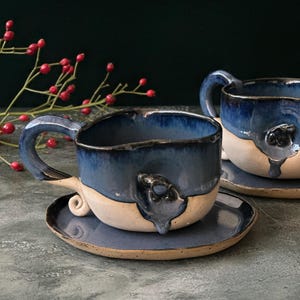 May include: Two handmade ceramic coffee mugs with blue and white glaze. The mugs have a unique, dripping glaze effect and are sitting on matching saucers.