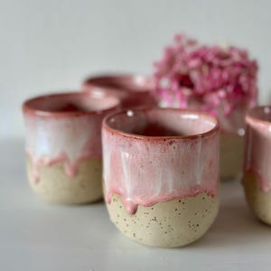 May include: A set of ceramic cups featuring a two-tone design with a speckled beige base and a glossy pink glaze that drips down from the rim. The cups have a rounded, handleless shape and a smooth texture on the upper portion.