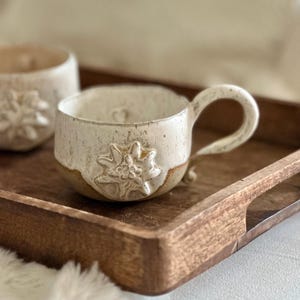 Könnte beinhalten: Zwei weiße Keramikbecher mit brauner Glasur am Boden. Beide Becher haben ein erhabenes weißes Blumendesign. Die Becher stehen auf einem Holztablett.