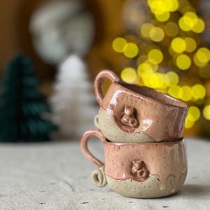 Op de afbeelding: Twee gestapelde keramische mokken met een gespikkeld glazuur in tinten bruin en roze. Elke mok heeft een reliëf ontwerp van een berenkop.