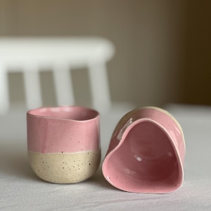 Op de afbeelding: Twee handgemaakte keramische bekers. Eén staat rechtop, de andere is gekanteld. Beide hebben een roze bovenste helft en een gespikkelde beige onderste helft. De bekers staan op een wit oppervlak, met een vage witte stoel op de achtergrond.