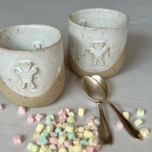Pode incluir: Duas canecas de cerâmica artesanais com um desenho de urso. As canecas são brancas com detalhes salpicados e uma base bege. Uma colher de prata repousa ao lado das canecas, e marshmallows coloridos estão espalhados.
