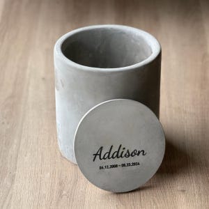 May include: A gray concrete container with a lid. The lid has the name "Addison" and the dates "06.12.2009 - 08.23.2024" engraved on it.