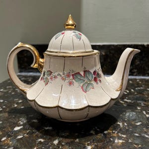 May include: A white teapot with a gold trim and floral design. The teapot has a lid with a gold finial and a spout on the side. The teapot is sitting on a dark brown surface.