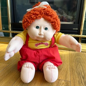 May include: A vintage Cabbage Patch Kids doll with orange yarn hair and a white bow. The doll wears a yellow shirt and red overalls. The doll's face is pale with green eyes, and the text "Cabbage Patch Kids" is visible on the shirt.