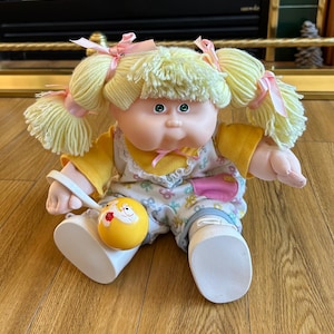 May include: A vintage Cabbage Patch Kids doll with blonde yarn pigtails tied with pink ribbons. The doll wears a yellow and floral patterned outfit, white shoes, and has green eyes. A yellow rattle with a cartoon design rests on its lap.
