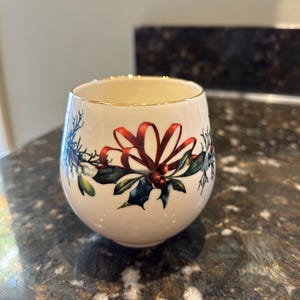 May include: A white ceramic cup with a gold rim and a festive holiday design featuring red bows, green holly leaves, and red berries.