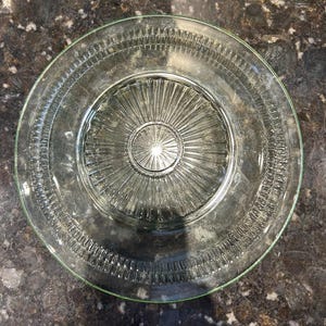 May include: A clear glass plate with a decorative design. The plate features a central sunburst pattern and a detailed border. The rim has a subtle green tint. This vintage plate is suitable for serving food.