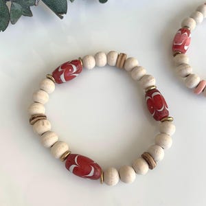 May include: Two beaded bracelets. Each bracelet features light wood beads, red and white patterned beads, and small gold-colored spacers. One bracelet is in focus, the other is slightly blurred in the background.