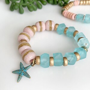 May include: A beaded bracelet with pink and gold beads, and light blue, irregular-shaped beads. A blue and gold starfish charm hangs from the bracelet. Another bracelet with pink and gold beads and light blue beads is in the background.