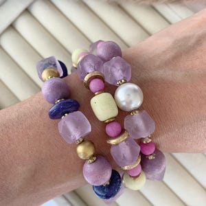 May include: Three beaded bracelets with a variety of beads in shades of purple, pink, and gold. The bracelets feature large, irregular-shaped beads, round beads, and a pearl. The bracelets are stacked together.