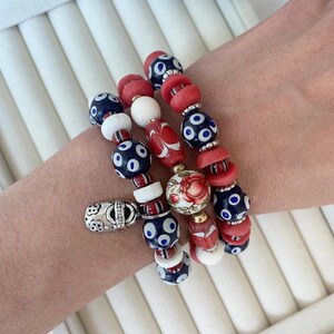 May include: Three beaded bracelets with a variety of colorful beads. The bracelets feature red, white, and navy blue beads with a floral design. One bracelet has a silver charm. The bracelets are on a white surface.