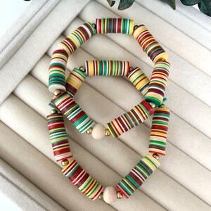 May include: Three colorful beaded bracelets. The bracelets are made of cylindrical beads in red, yellow, green, and white stripes. The bracelets are connected with wooden beads and gold-colored metal beads. The bracelets are displayed on a white surface.