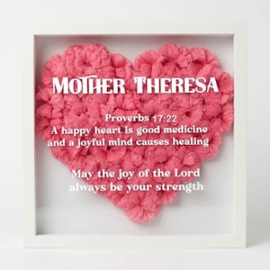 May include: A white shadow box frame containing a heart-shaped arrangement of pink carnations. The text reads: MOTHER THERESA, Proverbs 17:22, A happy heart is good medicine and a joyful mind causes healing, May the joy of the Lord always be your strength.