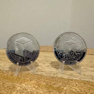May include: Two silver-colored coins with a 3D cube design. The coin on the left has the word "NEO" and the text "Digital Assets & Smart Contracts" around the edge. The coin on the right has the text "Smart Economy" and "2018" around the edge.