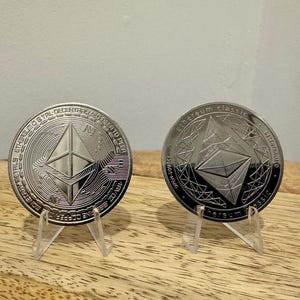 May include: Two silver Ethereum cryptocurrency coins on display stands. The coins feature the Ethereum logo and the text "Ethereum Classic" and "ETHEREUM is a TRULY DECENTRALIZED PLATFORM TO HELP ENABLE A GLOBAL, SECURE, AND OPEN FINANCIAL SYSTEM".