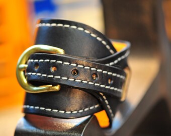 May include: A black leather strap with white stitching and a gold buckle. The strap is folded over and the buckle is closed.