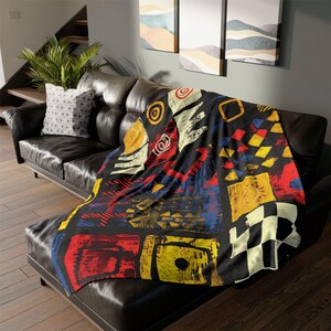 May include: A black throw blanket with a colorful abstract design featuring red, yellow, blue, and white shapes. The blanket is draped over a black leather sofa, adding a pop of color to the room.
