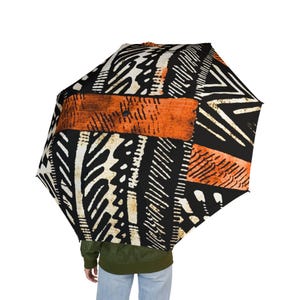May include: An open umbrella with a bold, geometric pattern in black, white, and orange. The design features abstract shapes and lines, creating a striking visual contrast. The umbrella is held by a person wearing a green jacket and blue jeans.