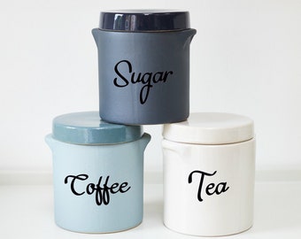 May include: Three ceramic canisters with lids, labeled "Sugar", "Coffee", and "Tea". The canisters are in shades of blue, gray, and white.
