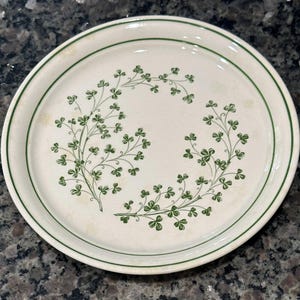 May include: A round, off-white plate with a green border and a decorative shamrock design. The shamrocks are arranged in a symmetrical pattern around the plate's center. The plate is approximately 9 inches in diameter.