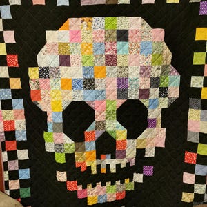 May include: A black quilt featuring a patchwork skull design. The skull is made from colourful fabric squares with floral and geometric patterns. The quilt is edged with a checkered border.
