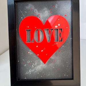 May include: Framed artwork with a red heart cut-out spelling "LOVE" against a grey, splattered background. The black frame surrounds the image. The artwork is suitable for home decor.