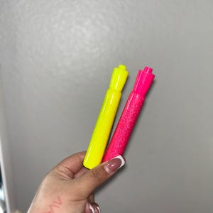 May include: Two glittery markers, one neon yellow and one hot pink, are held in a hand. The markers have a cylindrical shape with matching colored caps. The background is a plain, light gray wall.